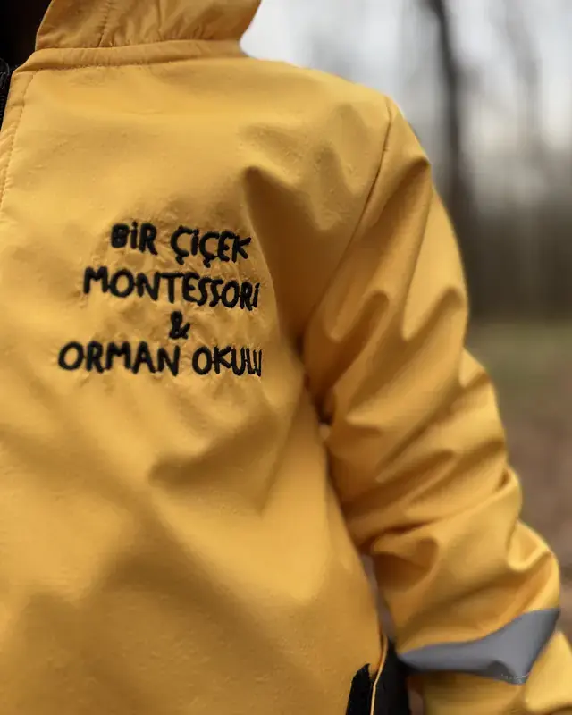 Sarı yağmurluk giymiş bir çocuk; montun üzerinde Bir Çiçek Montessori ve Orman Okulu yazıyor